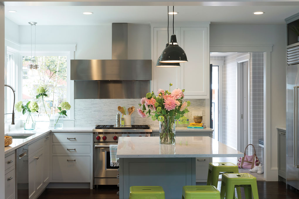 White cabinets surround a silver range hood accented by a marble mosaic. The island is surrounded by lime green stools, and a vase full of pink flowers sits on top.