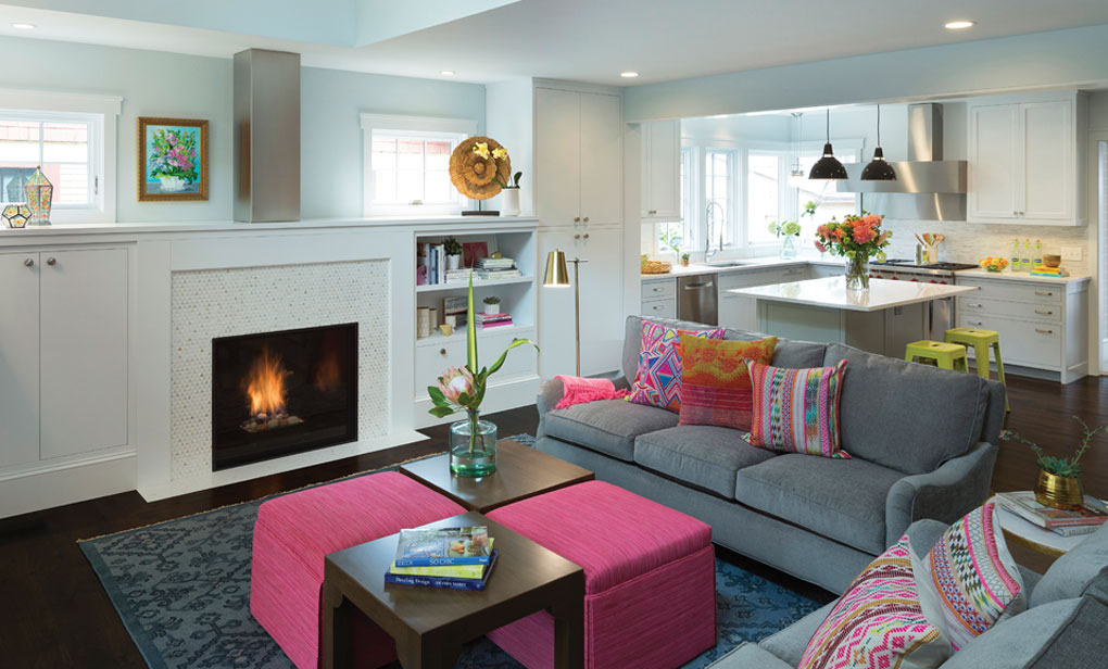 An open living room with fireplace is populated by gray couches and vibrantly-colored pillows. A a modern kitchen with clean lines can be found in the background.
