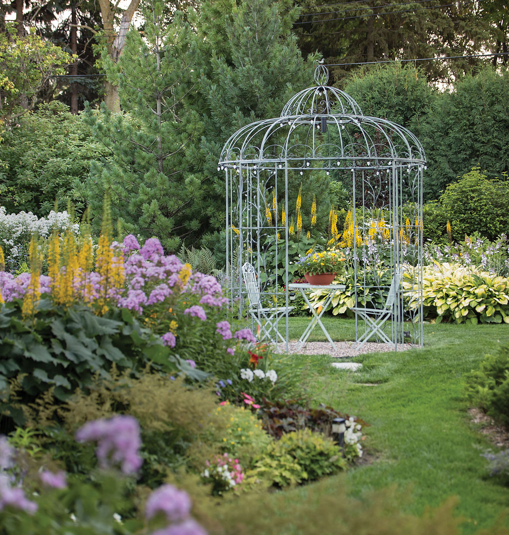 An open gazebo is surrounded by a lush garden full of green plants and colored flowers.