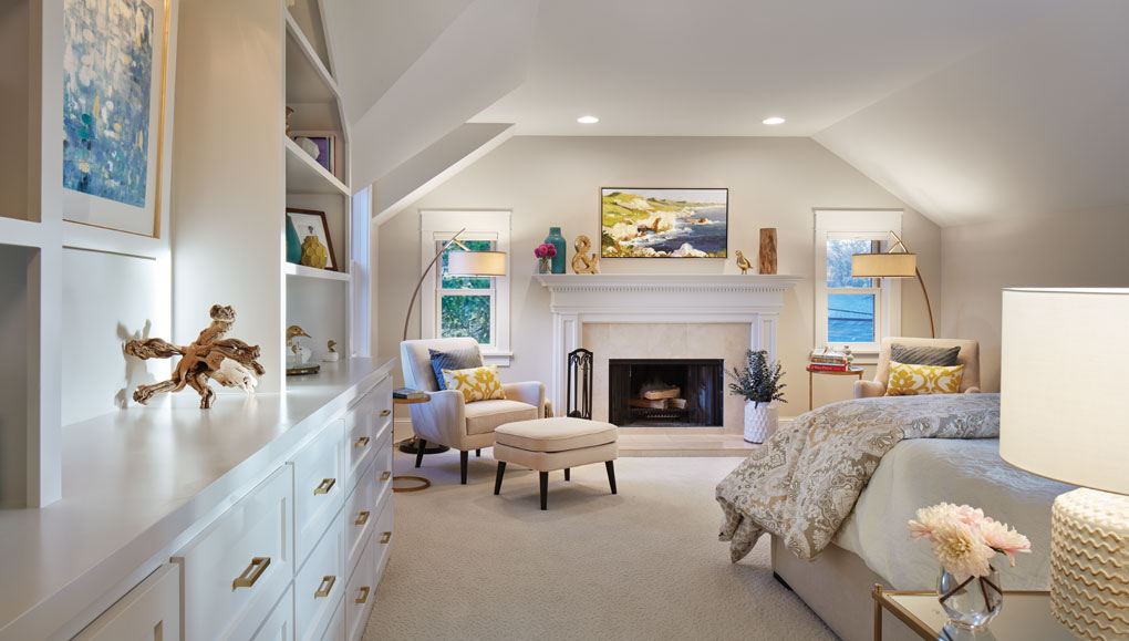 A master bedroom features white bookshelves, fireplace, and chair with accompanying footstool.