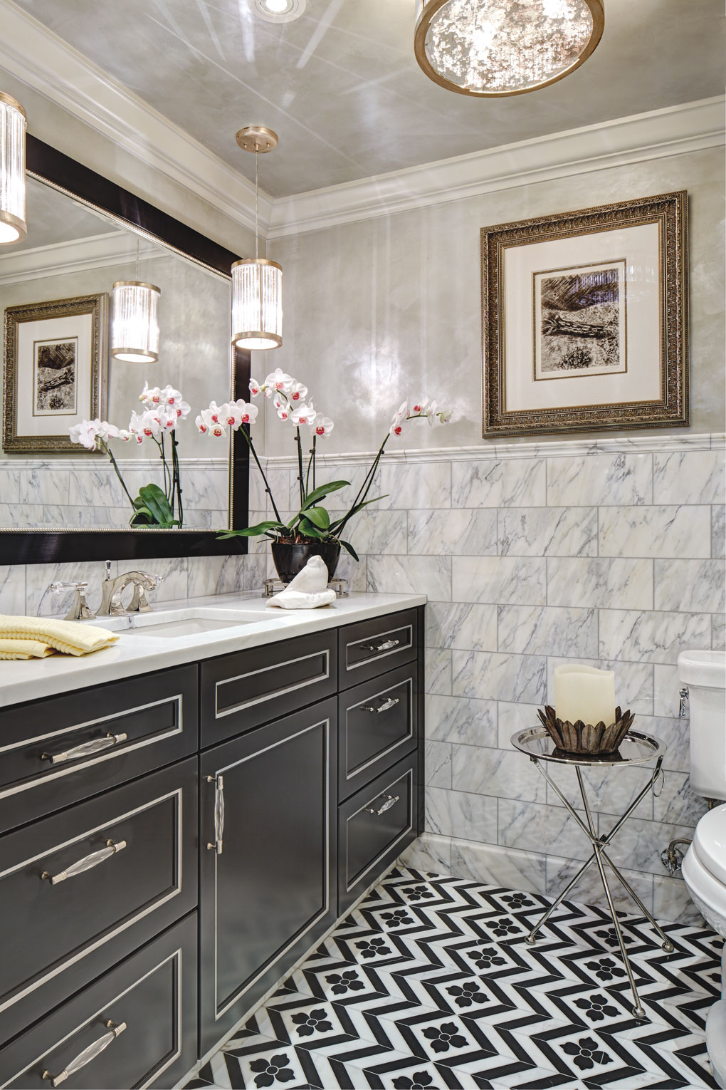 A bathroom with a sophisticated neutral color palette features tiled floor, marble walls, and chandeliers over the vanity.