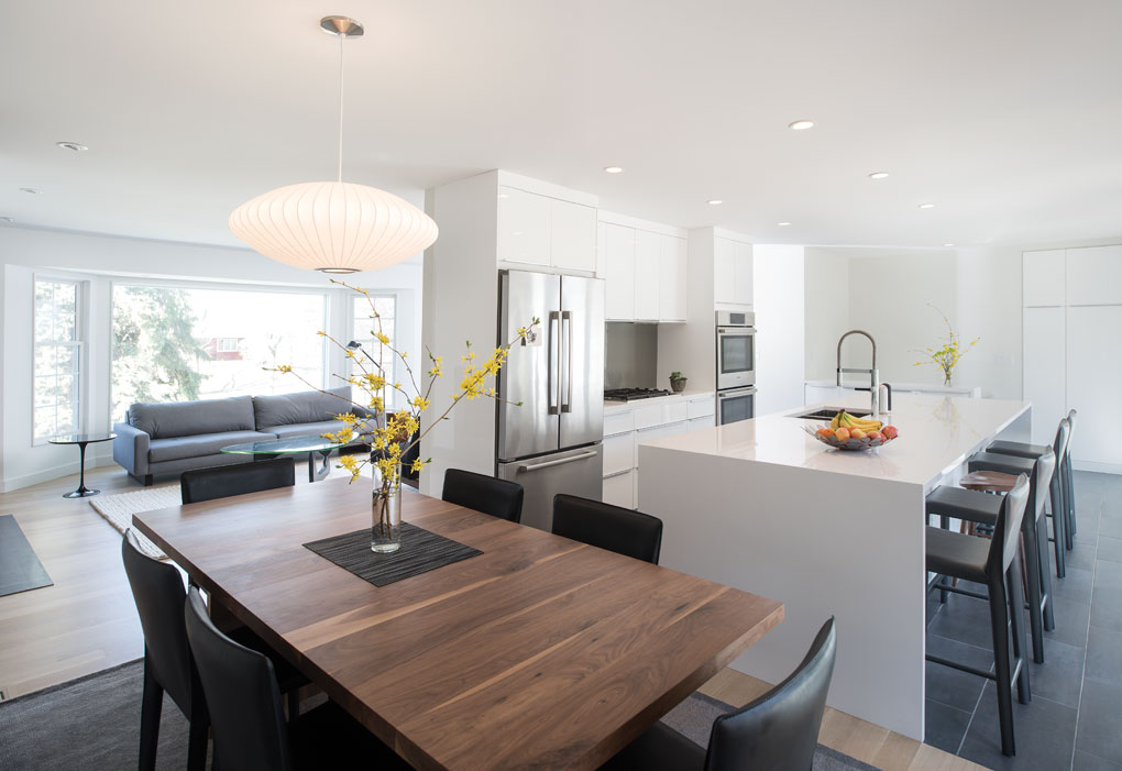 A sleek and modern kitchen is highlighted by a white, central island, white cabinetry and stainless steel appliances. A dark wood dining room table is adjacent to the island and is surrounded by black chairs.