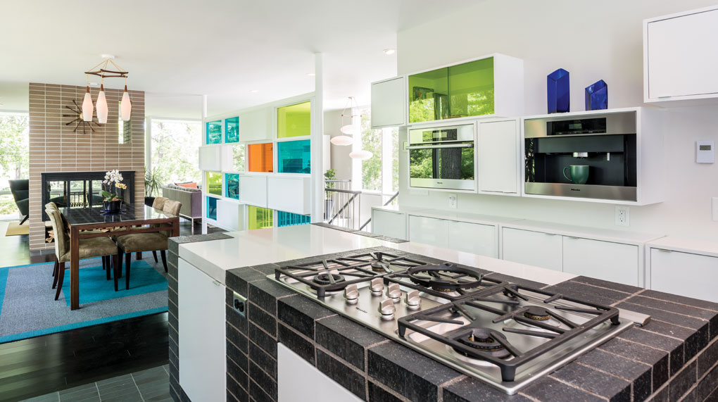 An update kitchen features an island made of stacked brick, white cabinetry and stainless steel appliances. A living room complete with fireplace is in the background.