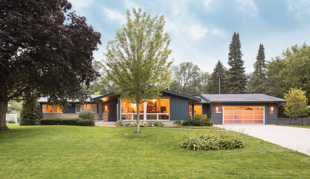 A blue rambler features large bay windows, and a garage door made of non-transparent glass. A large front yard with trees and shrubs is in the foreground.