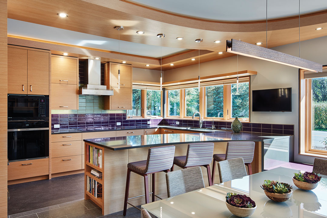 A kitchen designed by LiLu Interiors shows a center island with high chairs on one side, light wood cabinetry, tile backsplash, mounted television, and dining table.