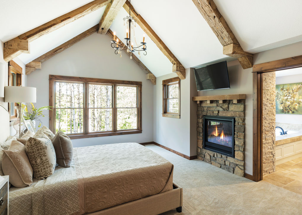 A master bedroom with a vaulted ceiling lined with exposed timber beams overlooks a stone fireplace and large bed.