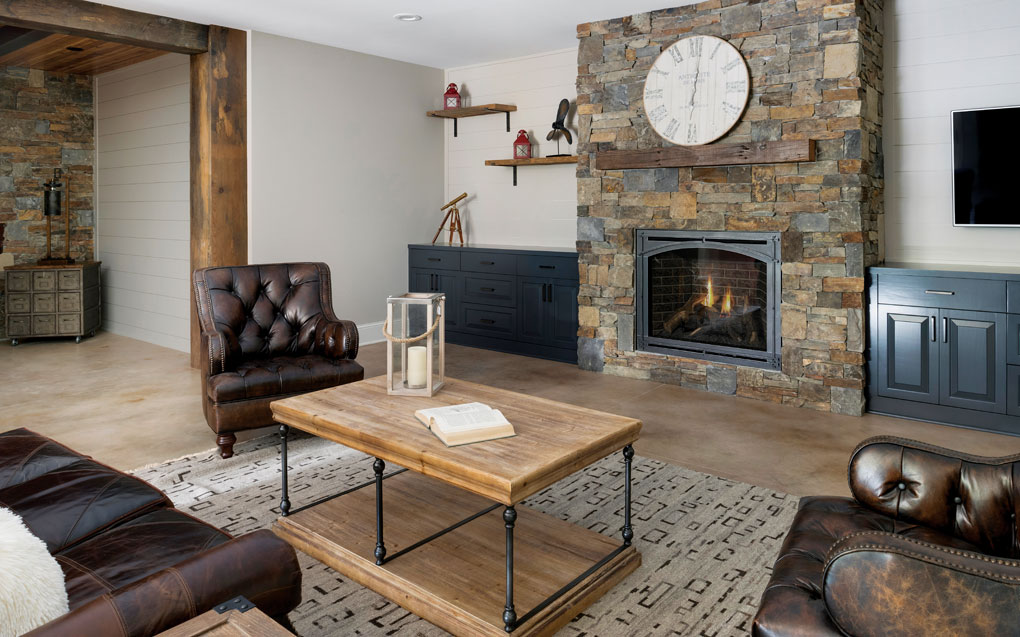 A basement is furnished with a leather sofa and chairs surrounding a coffee table that are set next to a large floor-to-ceiling, stone fireplace.