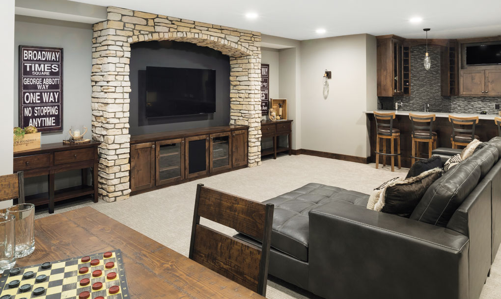A basement with a bar and leather sofa are set beside a stone arch housing a mounted television.