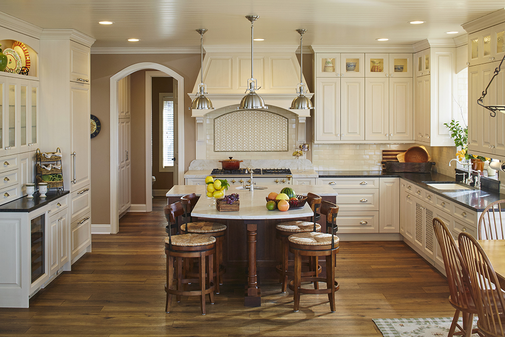 An ornate kitchen with cream-colored cabinetry, large range hood, hardwood flooring, center island with surrounding stools, and stone countertops.