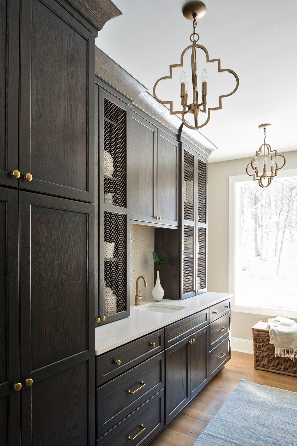 A room with large dark-wood cabinetry, two chandeliers, hardwood flooring, and a basket sitting next to a large window.