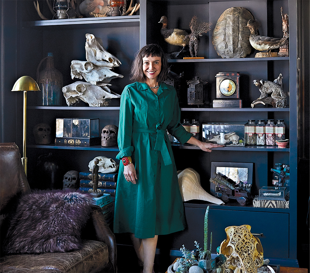 A photo of Christine Ward, owner of Patina, standing next to a shelf full of taxidermy and other collectibles in her home in a green dress.