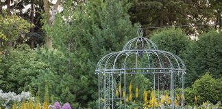 An open gazebo is surrounded by a lush garden full of green plants and colored flowers.