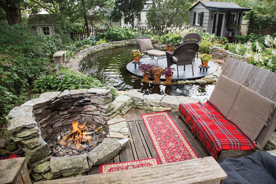 An island-like seating area set within the 3,000-gallon pond and a stone fire pit complete with its own cozy pallet seating offer additional spaces to relax in this luxurious backyard oasis.