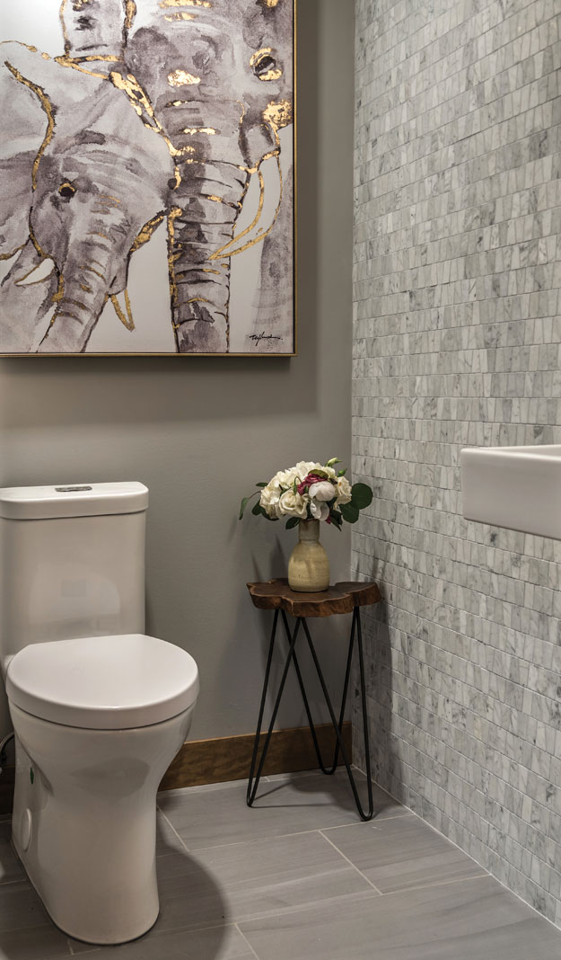 A small, renovated bathroom that features an accent wall made from mosaic Carara marble tiles, small table with with a vase of flowers on top, toilet, and elephant painting.